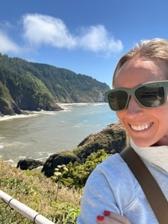 Woman wearing sunglasses and a light gray hoodie smiles for a selfie on a cliffside trail with a rocky coastline and blue sky behind her.