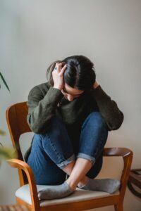 Person seated on a wooden chair, hunched over with hands on their head in distress or frustration.
