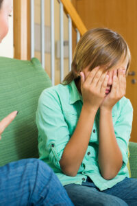 Girl sitting on a green couch, hands covering her face in distress during a conversation with an adult nearby.