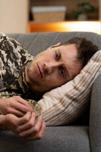 Man in camouflage clothing lying on a gray sofa with his head resting on a striped pillow, looking tired or at ease.