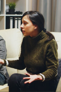 Woman with dark hair in a green turtleneck sits on a cream sofa, mid-conversation, hands gesturing.